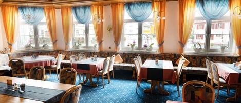 A restaurant with multiple tables and chairs, large windows with curtains, and a patterned carpet.
