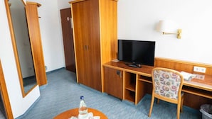 A hotel room with a wooden wardrobe, a desk with a television, a chair, and a small table with a bottle and glasses.