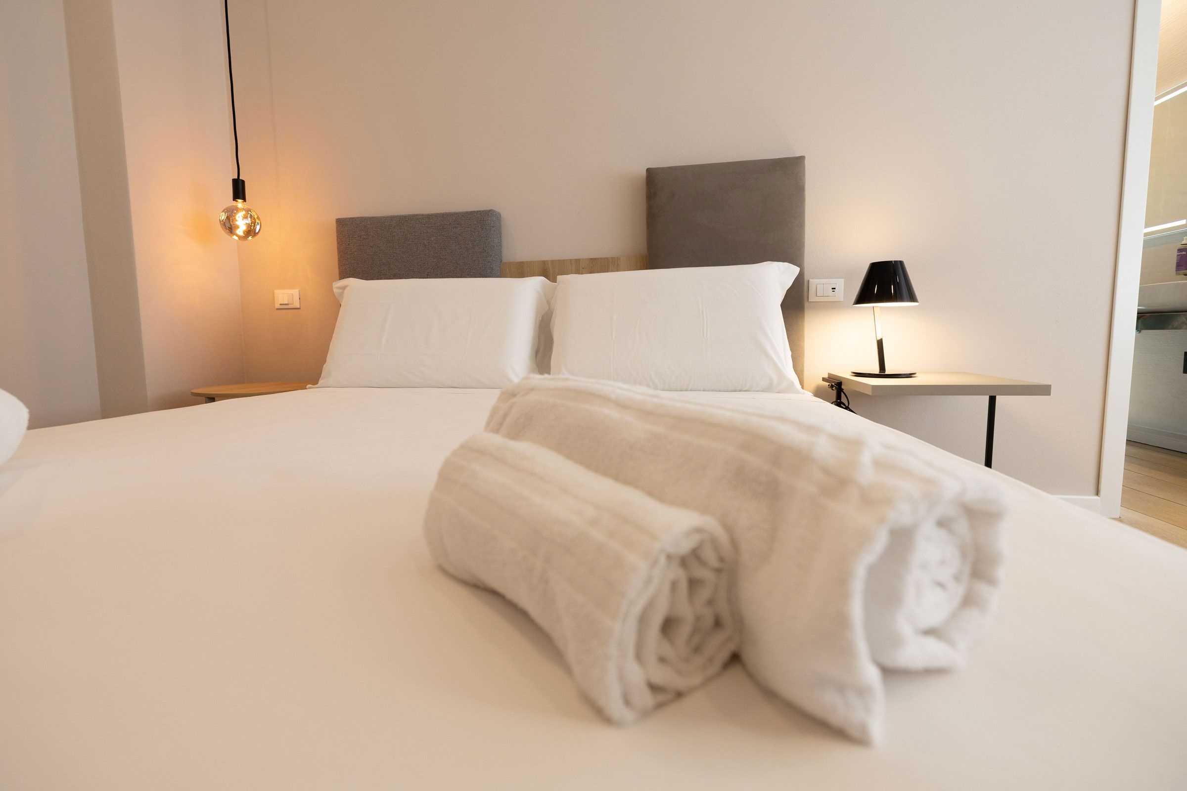 Un lit bien fait, avec une serviette enroulée, une lampe de chevet et un luminaire fixé au mur, dans une chambre d’hôtel.