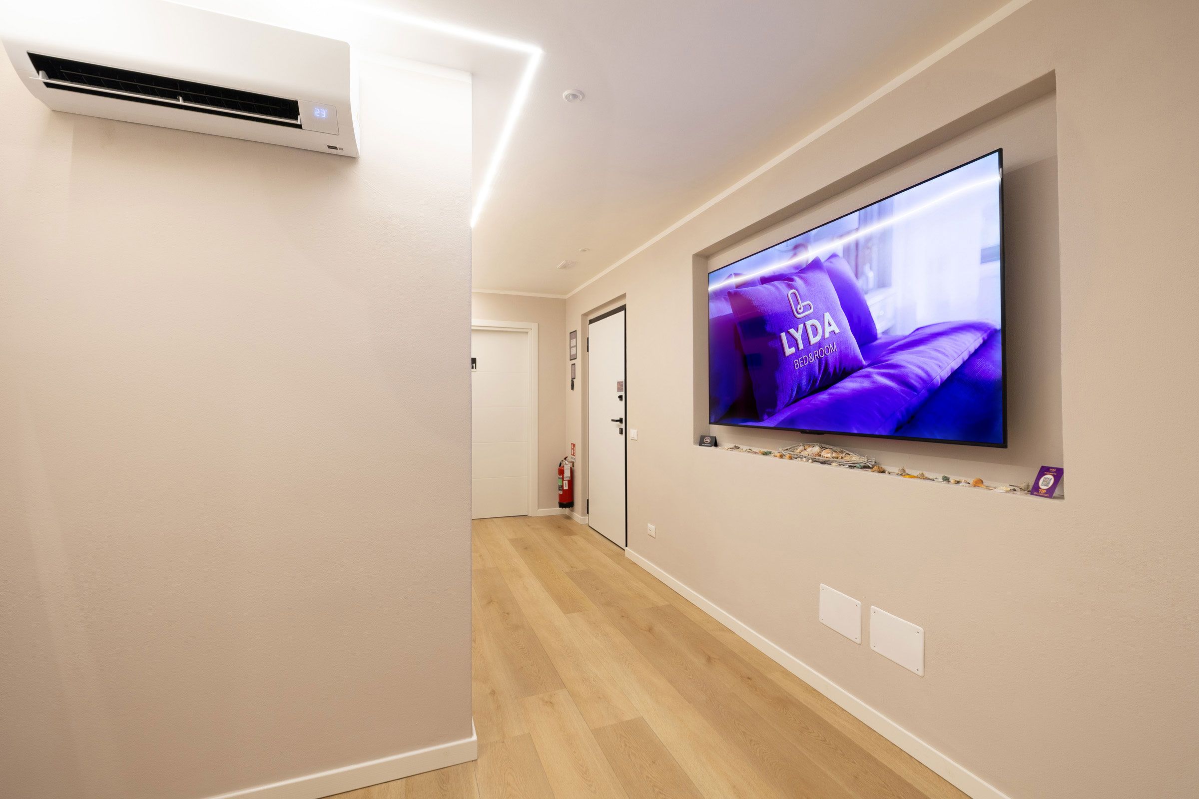 Un pasillo con un televisor de pantalla plana montado en la pared que muestra un anuncio con temática púrpura, una unidad de aire acondicionado blanca y un extintor.
