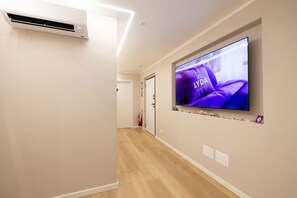 Un pasillo con un televisor de pantalla plana montado en la pared que muestra un anuncio con temática púrpura, una unidad de aire acondicionado blanca y un extintor.