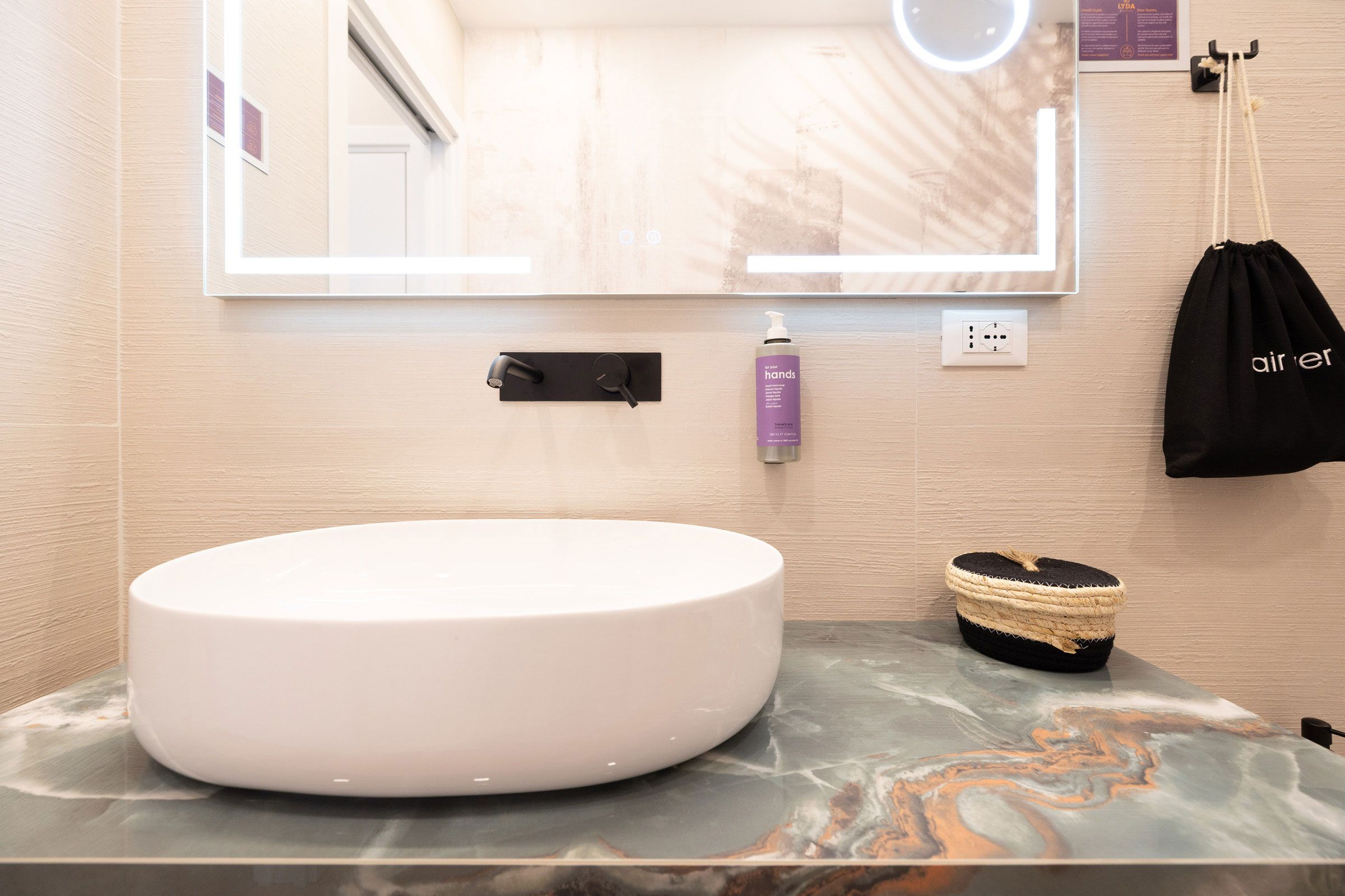 Une salle de bain avec un lavabo rond blanc, un miroir, un distributeur de serviettes noir, un sac noir et un panier en osier.