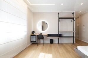 A modern hotel room with a wooden floor, a desk with a chair, a small refrigerator, a round mirror, a black shelf unit, and a bed. - Lyda Bed&Room (La Spezia)