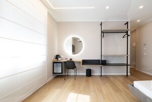 A modern hotel room with a wooden floor, a desk with a chair, a small refrigerator, a round mirror, a black shelf unit, and a bed.