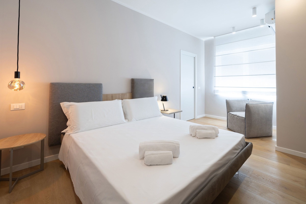 Habitación de hotel moderna con una cama grande, mesitas de noche, un escritorio, una silla y una ventana con persianas.
