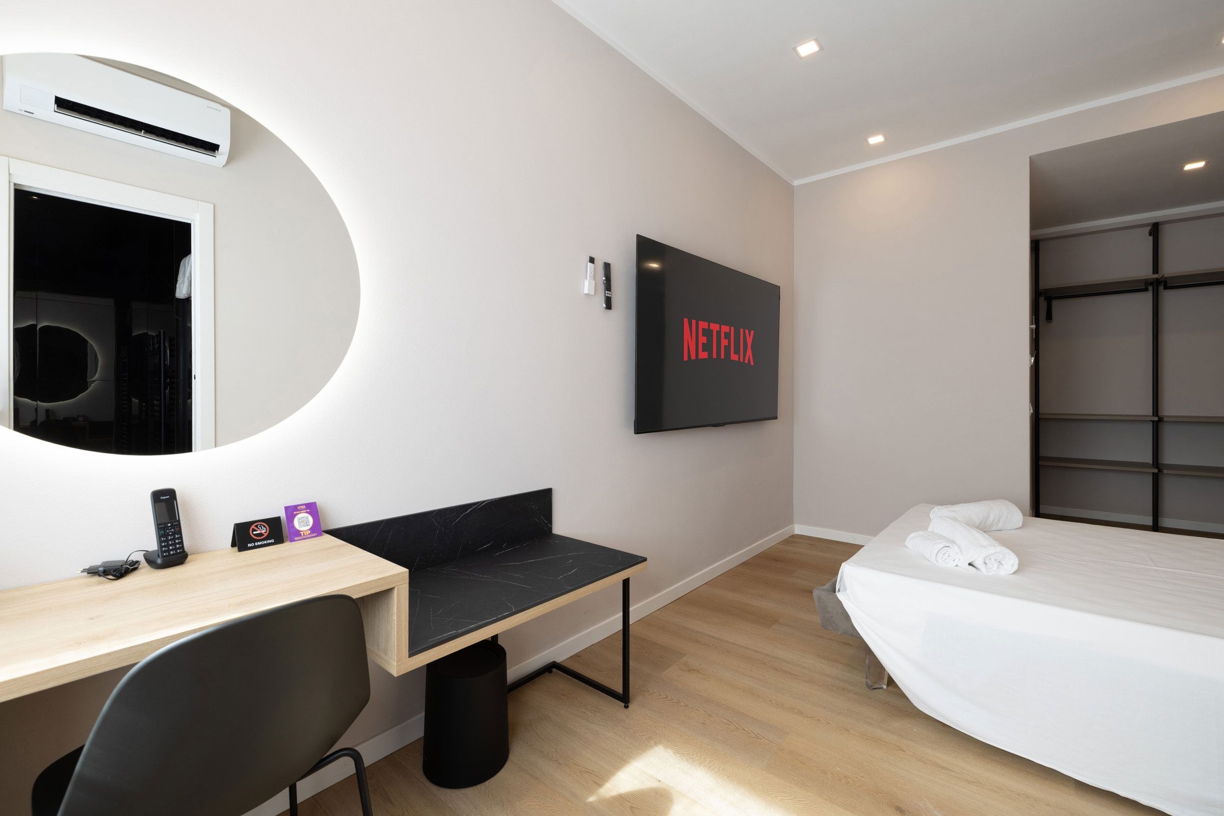 Una camera d'albergo con un letto, una scrivania con una sedia, un televisore a parete, un cartello che indica il divieto di fumare e un cestino.