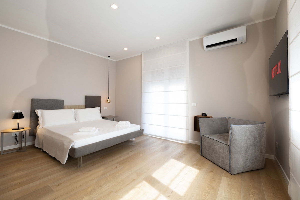 Una habitación de hotel moderna con cama, mesita de noche, lámpara, equipo de aire acondicionado y una zona de estar con una silla.