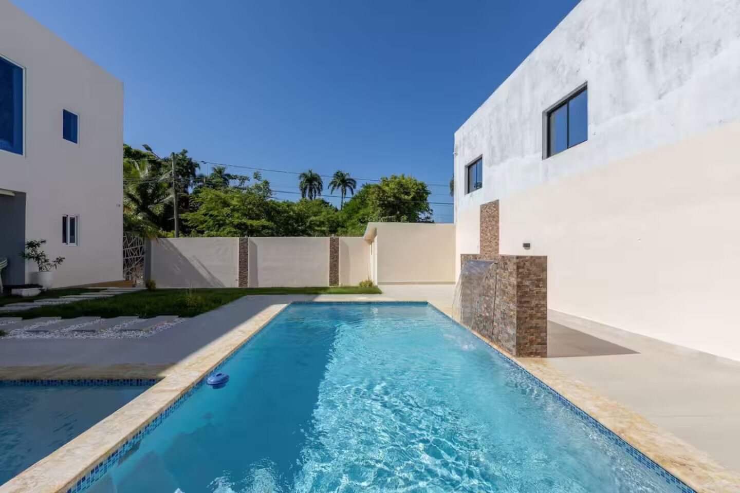 A rectangular swimming pool with a clear blue water surface, surrounded by a white building with a flat roof and a stone accent wall.