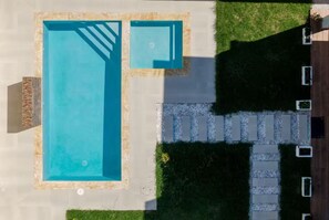 Ein Luftbild eines Schwimmbeckens mit umlaufender Sonnenterrasse, einem kleinen angrenzenden Pool und einem Weg zu einem Gebäude.