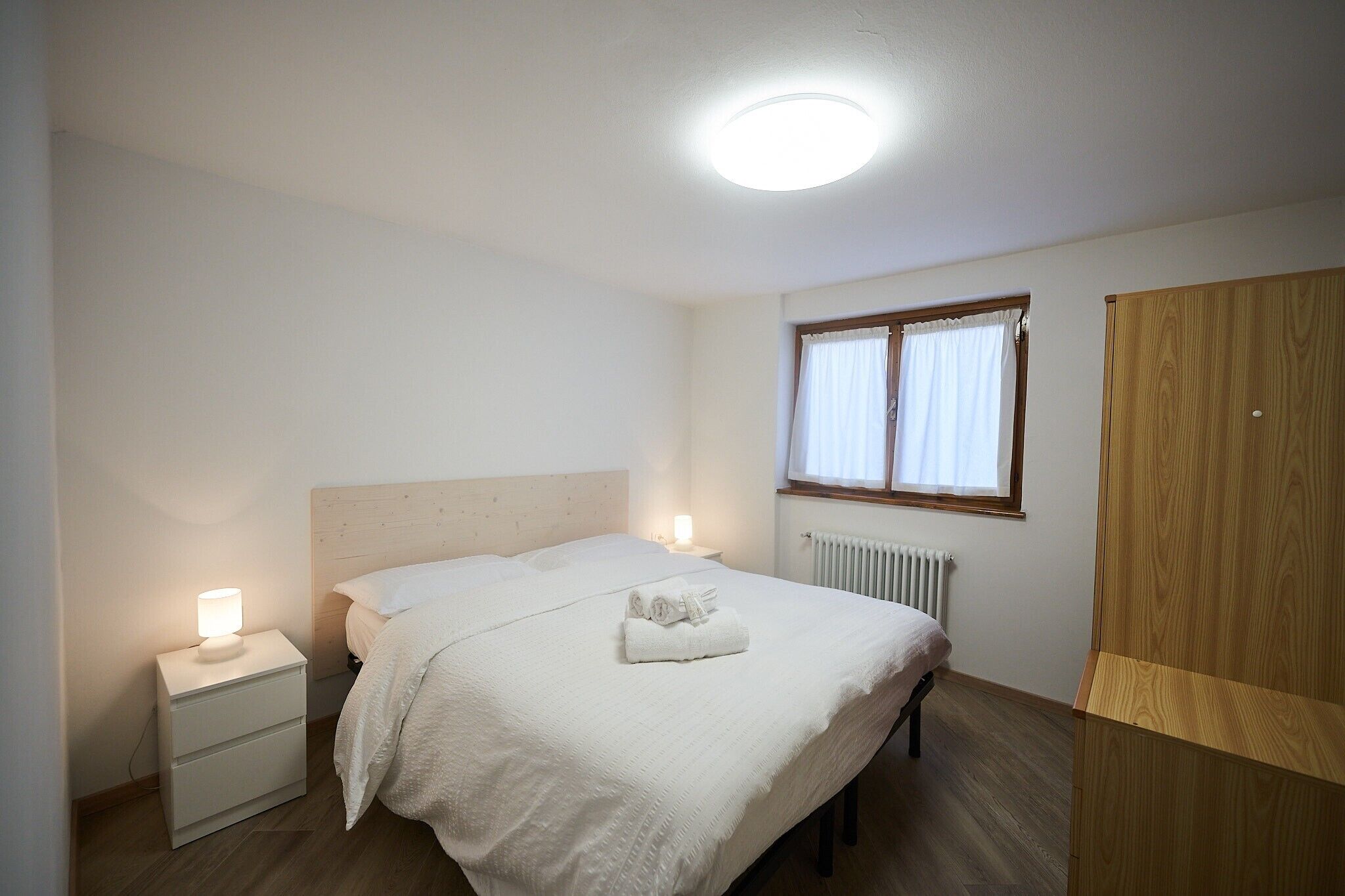 Et soveværelse med seng, sengeborde, et vindue med gardiner, en radiator og et klædeskab.