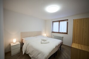 Ein Schlafzimmer mit Bett, Nachttischen, einem Fenster mit Vorhängen, einem Heizkörper und einem Kleiderschrank.