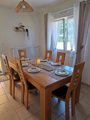 Zona de menjador amb taula de fusta preparada per a sis persones, envoltada de cadires, una làmpada penjant i una finestra amb cortines.