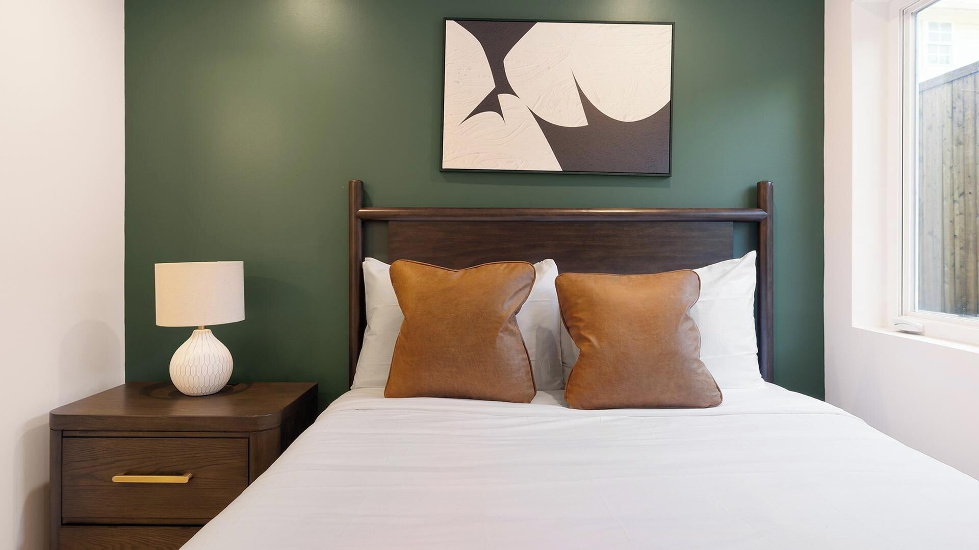 A neatly made bed with two pillows, a bedside table with a lamp, and a wall-mounted abstract artwork.