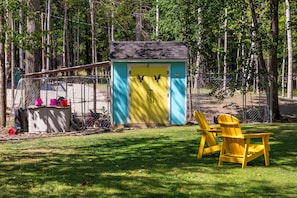 A small, colorful shed with a door, a fenced area with a chicken coop, and a yellow chair in a grassy yard. -  Welcome to The Crooked Road Cottage at Acadia! NO cleaning fees. NO pet fees. (Bar Harbor)