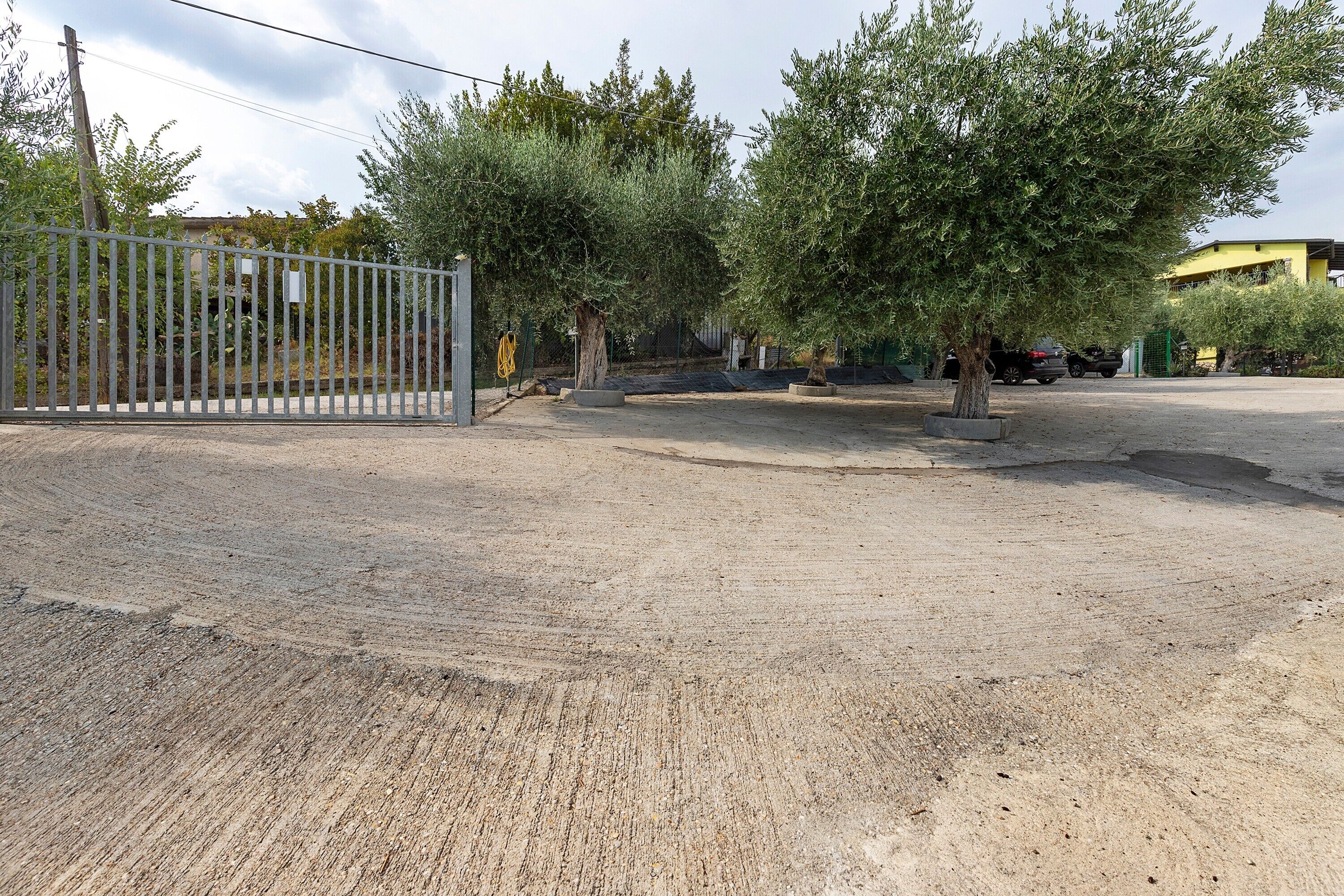 A gravel parking area with a metal gate, trees, and a building in the background.