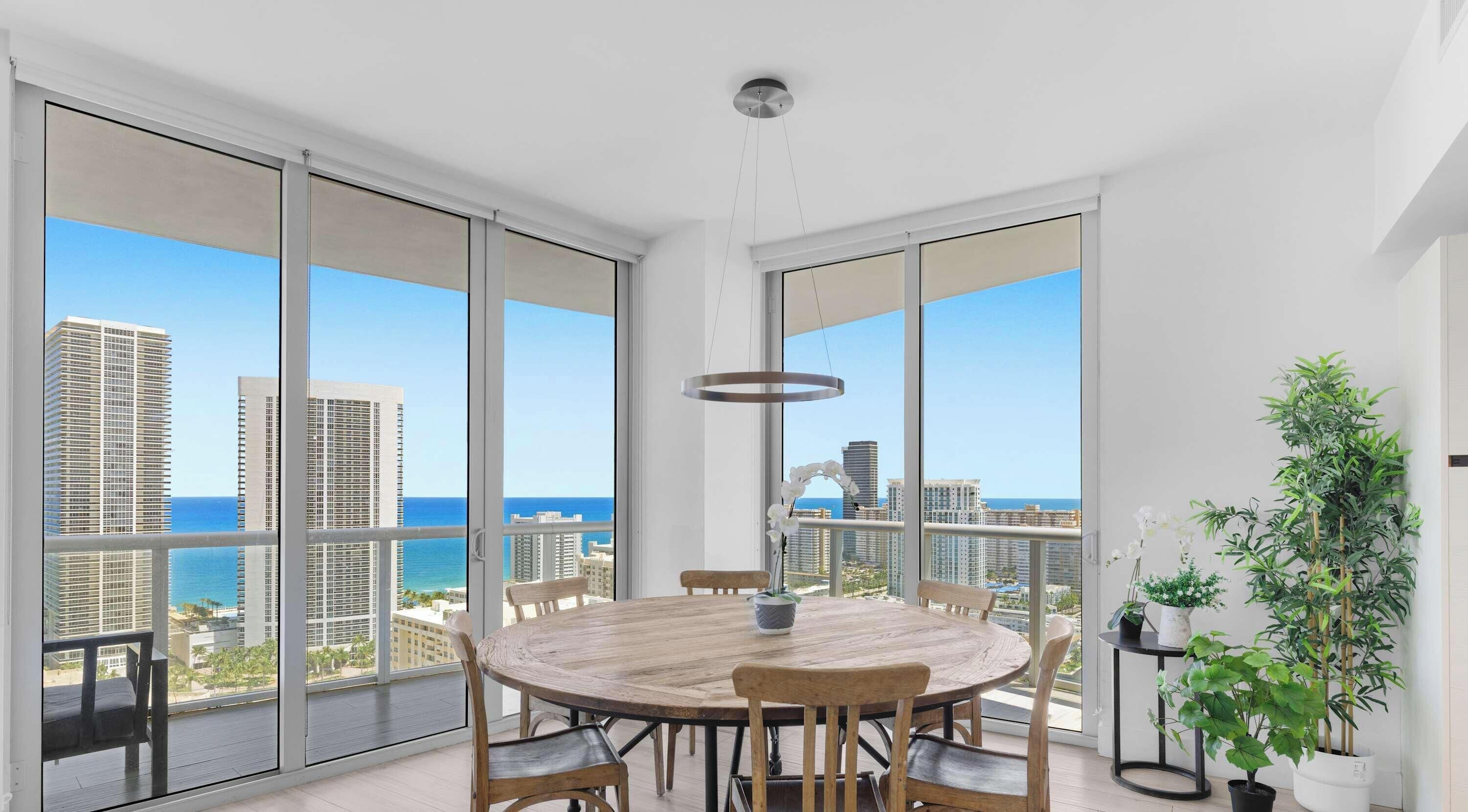 Een ruim balkon met een houten tafel en stoelen, grote ramen met uitzicht op de stad en de oceaan, en een hanglamp.