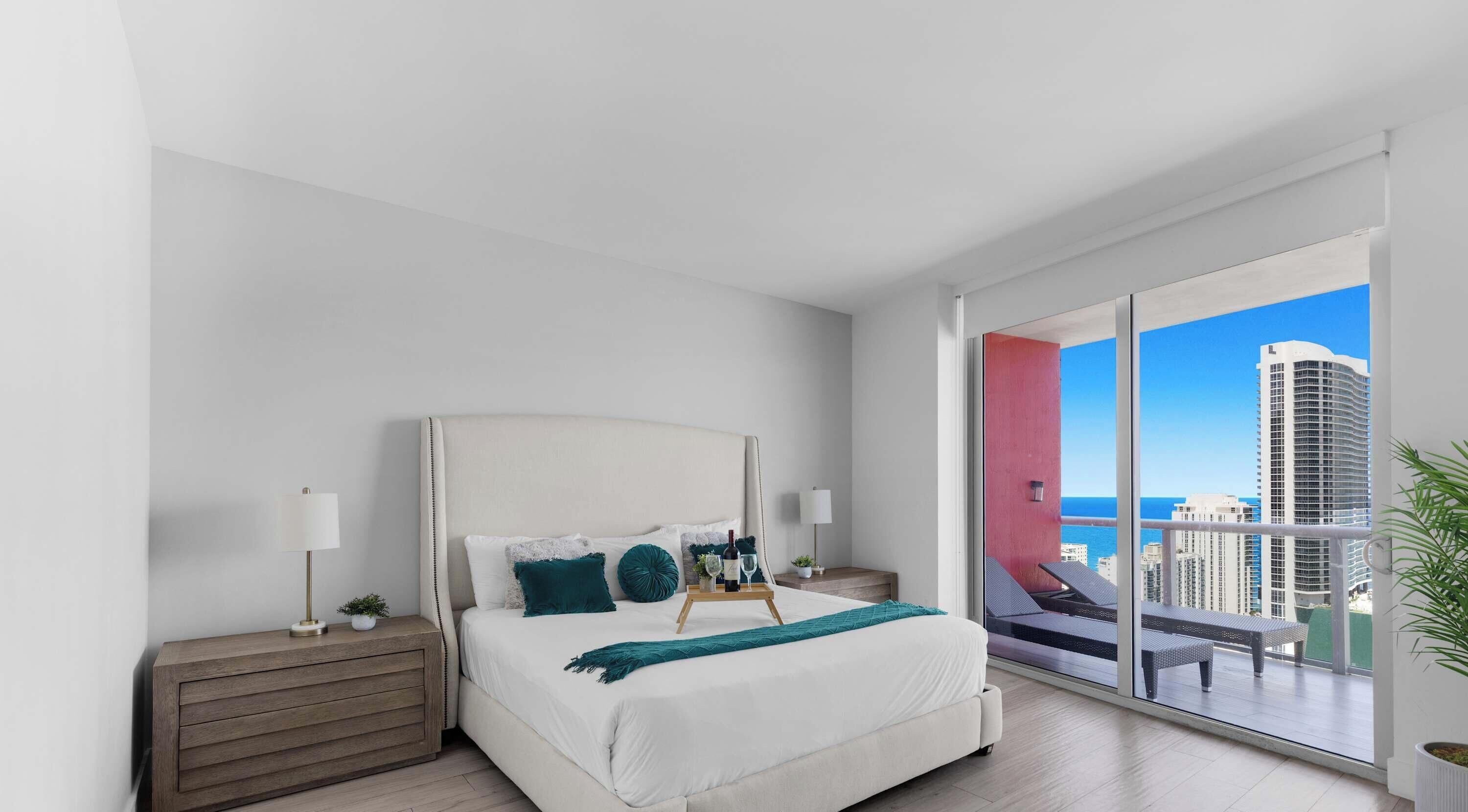 Une chambre moderne avec un grand lit, des tables de chevet, une commode et un balcon offrant une vue sur la ville et l’océan.