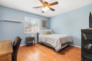 Ein Schlafzimmer mit Bett, Deckenventilator, Fenster mit Jalousien, Nachttisch und Sessel.