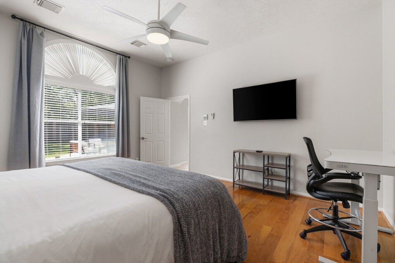Ein Schlafzimmer mit Bett, Schreibtisch, Stuhl, Fernseher und Deckenventilator.