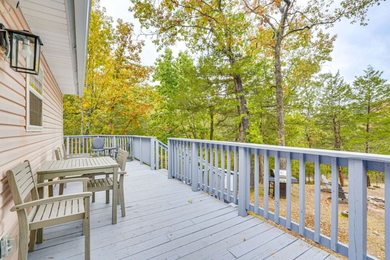 A wooden deck with a railing, a table, and chairs, surrounded by trees with autumn foliage.