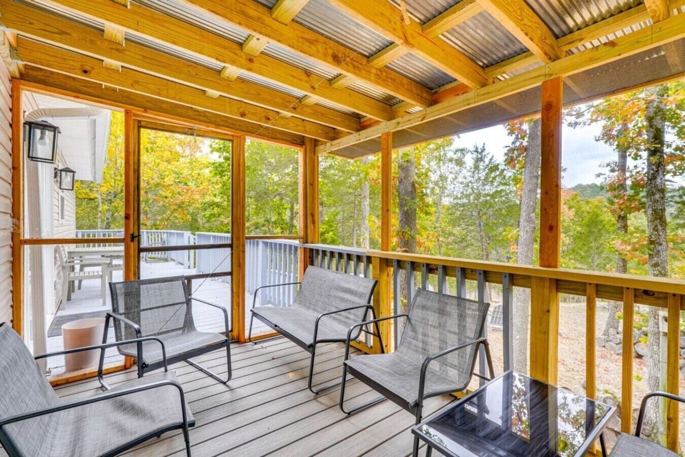 Une terrasse extérieure couverte, dotée de chaises à armature métallique et d’une table en verre, qui offre une vue sur une zone boisée.