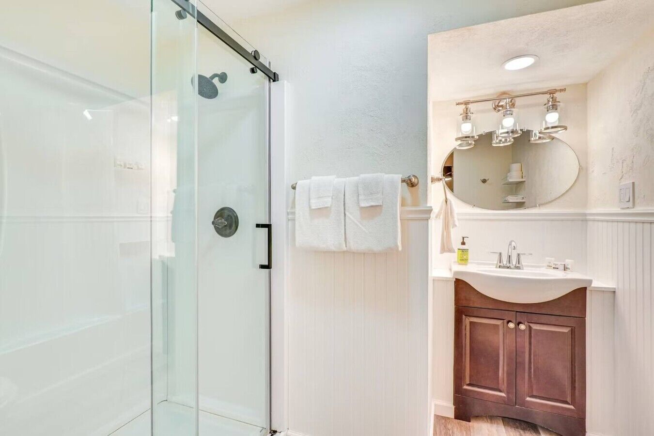 A bathroom with a glass shower enclosure, a white sink with a vanity, and a mirror with lights.