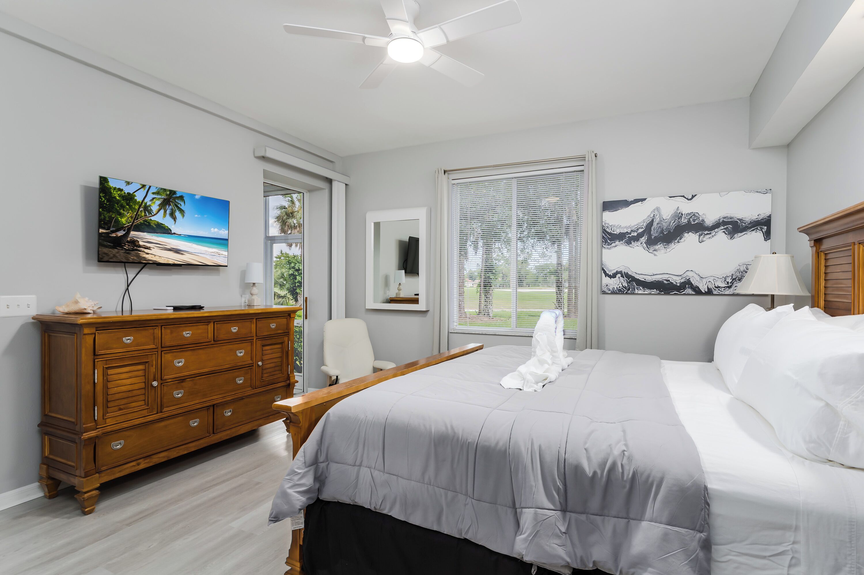 A bedroom with a bed, a wooden dresser, a television, a window with blinds, and a ceiling fan.
