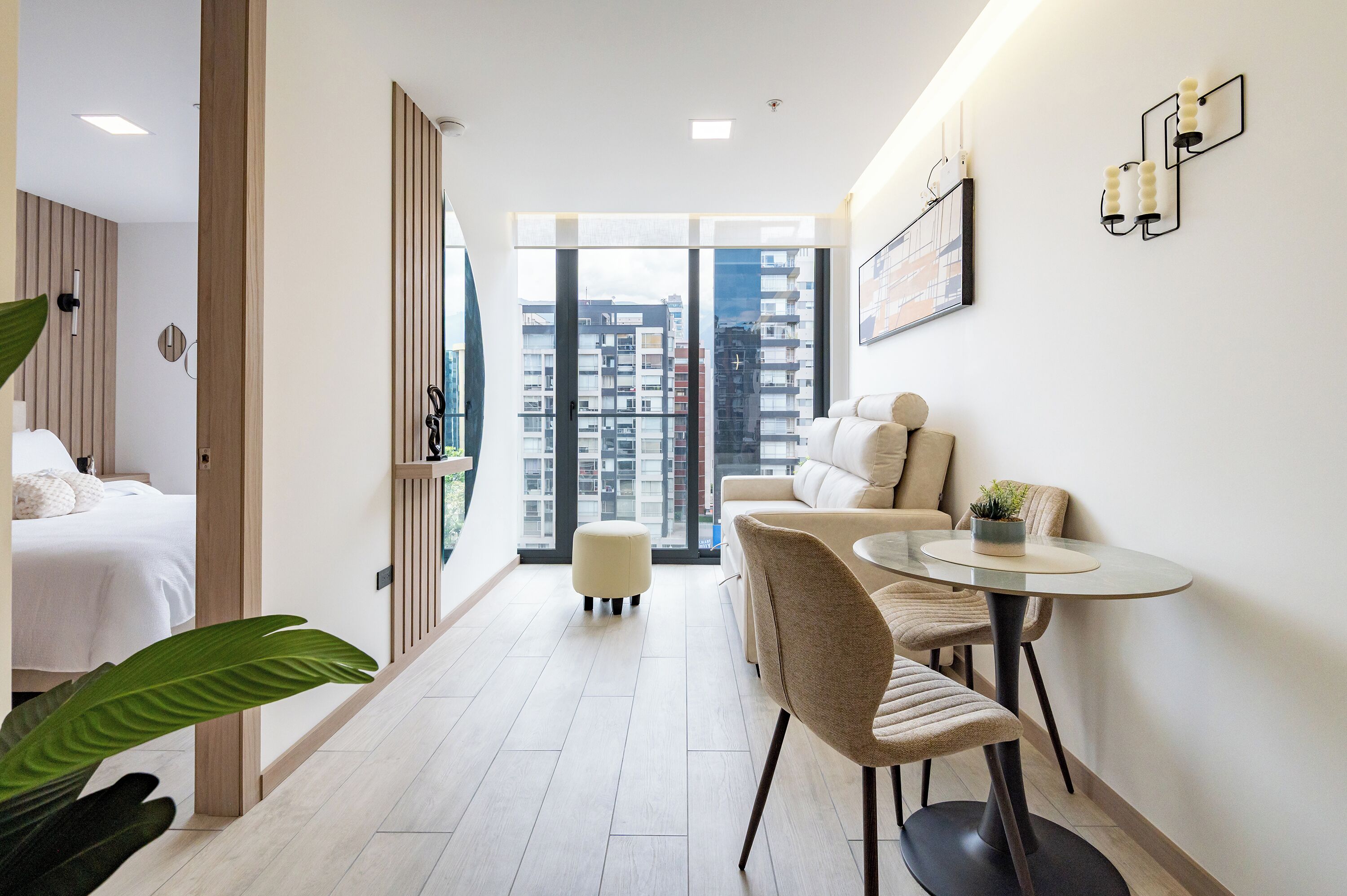 A modern hotel room with a bed, a seating area with a table and chairs, and a large window offering a city view.
