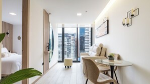 Et moderne hotelværelse med seng, siddeområde med bord og stole samt et stort vindue med udsigt over byen.