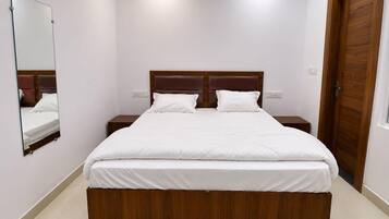 Une chambre à coucher avec un lit en bois, du linge de lit blanc, une table de chevet, un miroir et une porte en bois.