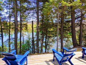 A wooden deck with blue chairs overlooking a lake surrounded by tall trees. - Ambre (Grenville-sur-la-Rouge)