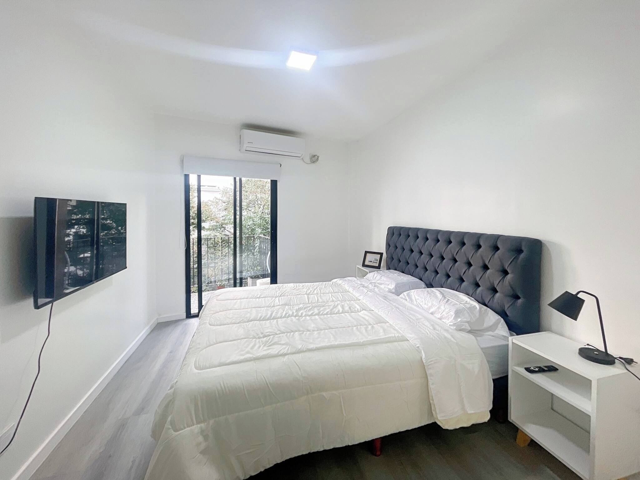 Une chambre à coucher moderne avec un grand lit, un téléviseur fixé au mur et une porte-fenêtre coulissante donnant sur l’extérieur.