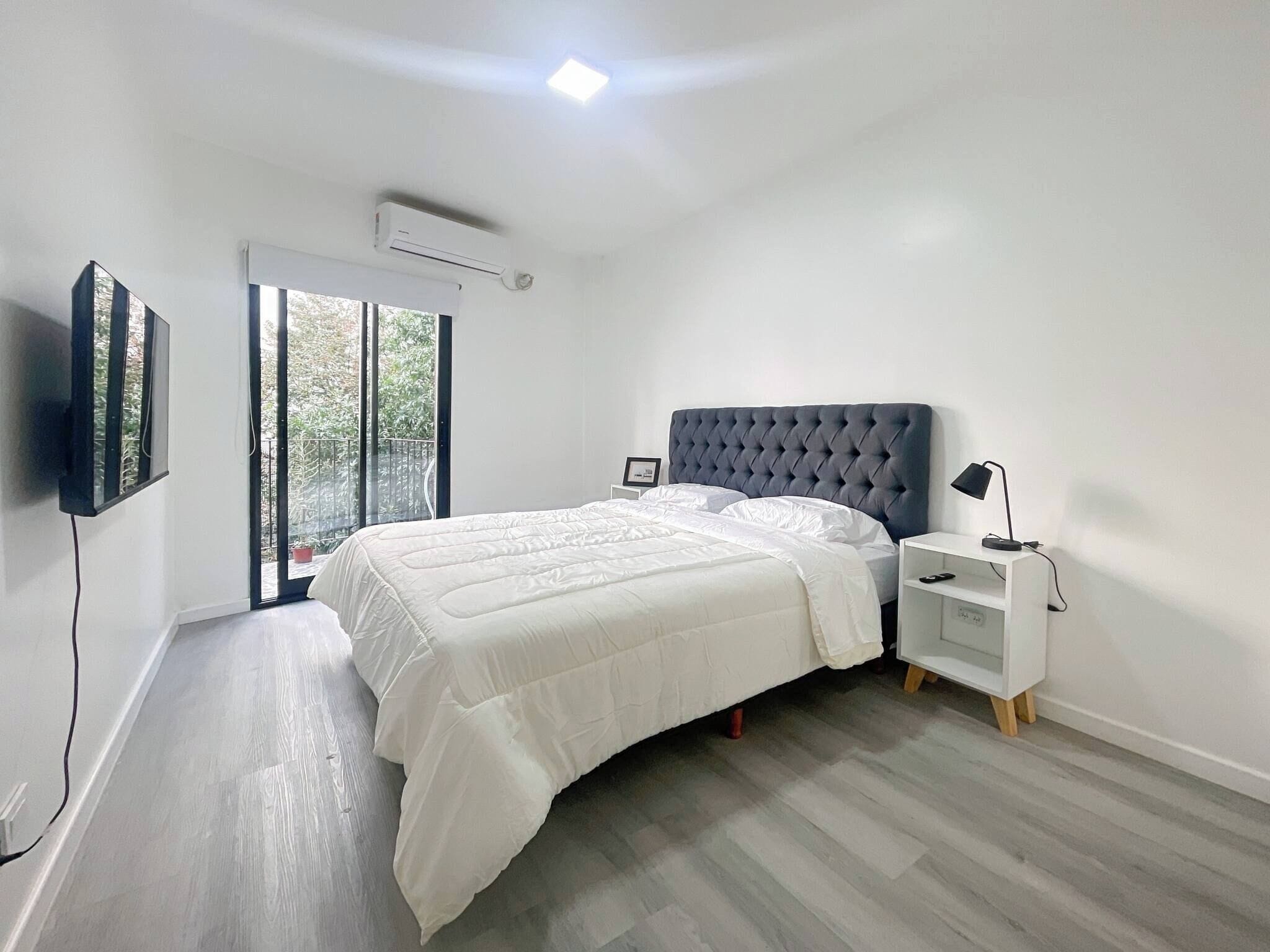 Une chambre à coucher moderne avec un grand lit, un téléviseur fixé au mur, une table de chevet avec une lampe et une porte-fenêtre coulissante donnant sur un balcon.