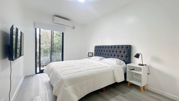 A modern bedroom with a large bed, a TV mounted on the wall, a bedside table with a lamp, and a sliding glass door leading to a balcony. - Charming Apartment in Recoleta 4 pax (Buenos Aires)