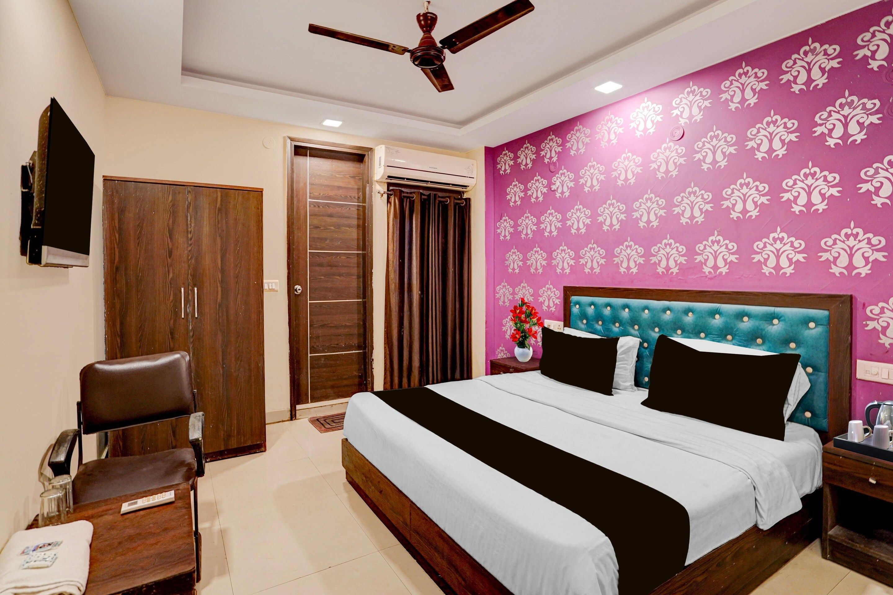 Une chambre d’hôtel avec un lit, une chaise, un bureau, un ventilateur de plafond et un mur orné d’un motif floral.