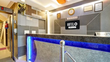 A hotel reception desk with a blue illuminated feature, a clock, framed certificates, and a sign indicating check-in times.