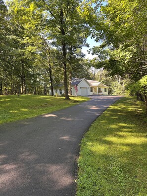 A paved pathway leading to a single-story building with a gabled roof, surrounded by lush green trees and grass.