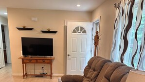 A living room with a sectional sofa, a flat-screen TV on a wooden stand, a door with a decorative glass panel, and a patterned curtain. - Fabulous 3-bedroom house on 2 acres in quiet Garrett County wooded neighborhood (Frostburg)