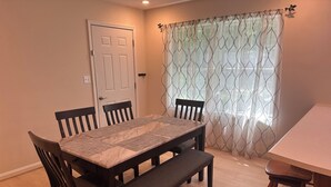 Un comedor con una mesa rectangular, cuatro sillas, un banco y una ventana con cortinas estampadas.