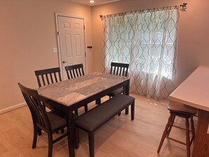 A dining area with a rectangular table, four chairs, a bench, and a window with patterned curtains.