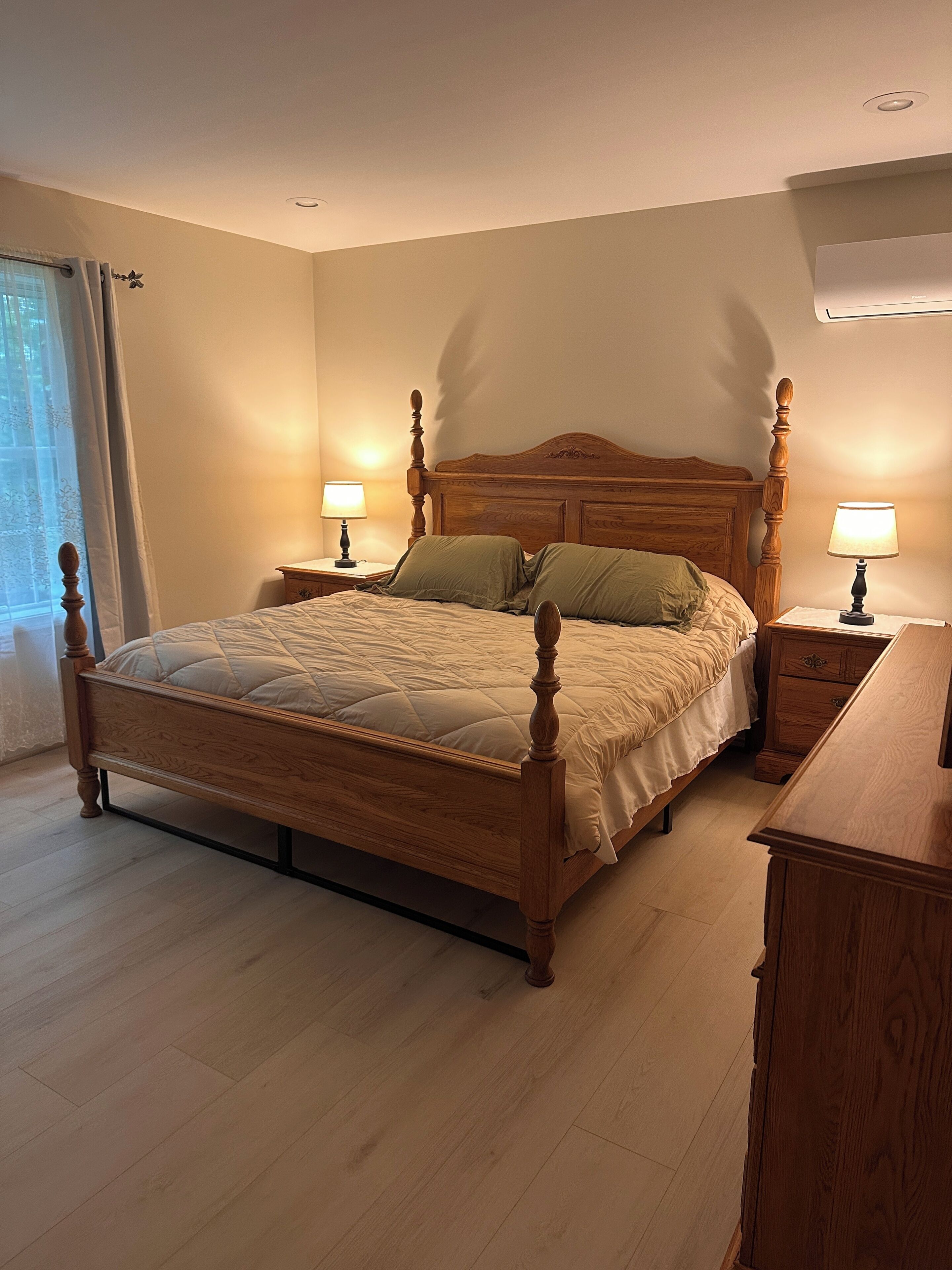 A bedroom with a wooden bed, two bedside tables with lamps, a wooden dresser, and a window with curtains.