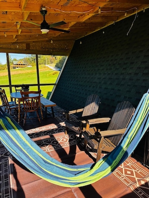 A hammock chair set on a patterned rug, wooden chairs, a table, and a view of a green landscape through a large window. - Newly built cabin by Luray Caverns/Shenandoah Natl Park/Wedding Venues (Stanley)