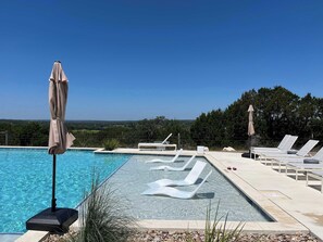 A clear blue swimming pool with lounge chairs and umbrellas, surrounded by a landscaped area with trees. - Firefly Luxury Rv Resort - Deluxe RV Site-324 (Fredericksburg)