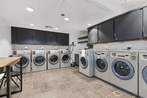 A laundry facility with multiple washing machines, a vacuum cleaner, and a wooden table. - Firefly Luxury Rv Resort - Deluxe RV Site-324 (Fredericksburg)