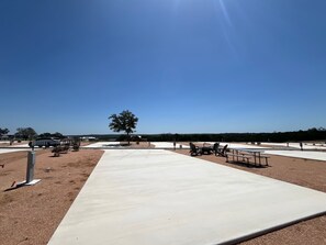 An outdoor area with a paved walkway, picnic tables, and a tree. - Firefly Luxury Rv Resort - Deluxe RV Site-324 (Fredericksburg)