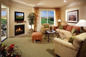 A living room with a fireplace, a television, a sofa, a coffee table, and a view of a golf course. - 1 BR Shadow Ridge Villa during Coachella Music Festival (Palm Desert)