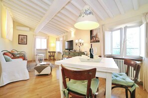 Ein Essbereich mit einem weißen Tisch, Stühlen und einer Deckenleuchte. Ein Fernseher steht auf einem Schrank, und es gibt ein Fenster mit Vorhängen.