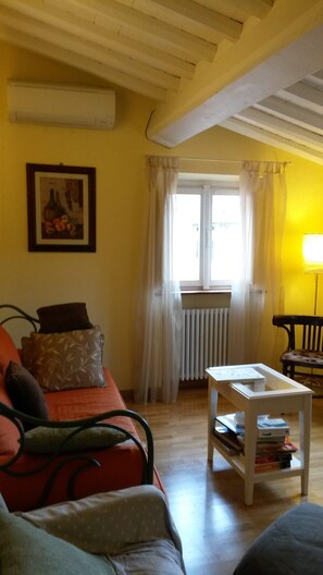 Ein Wohnzimmer mit einer Couch, einem Tisch mit Büchern, einem Heizkörper und einem Fenster mit Vorhängen.