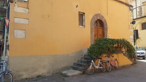 Ein Gebäude mit gelber Fassade, einer Holztür und einem kleinen Fenster. Vor dem Eingang führen Stufen hinauf, und ein Fahrrad steht davor abgestellt.