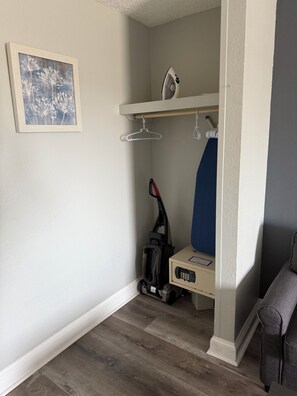 A corner of a room with a wooden floor, a white wall, a framed picture, a vacuum cleaner, and a blue chair. - Your Beachfront Getaway Awaits at Myrtle Beach SC (Myrtle Beach)